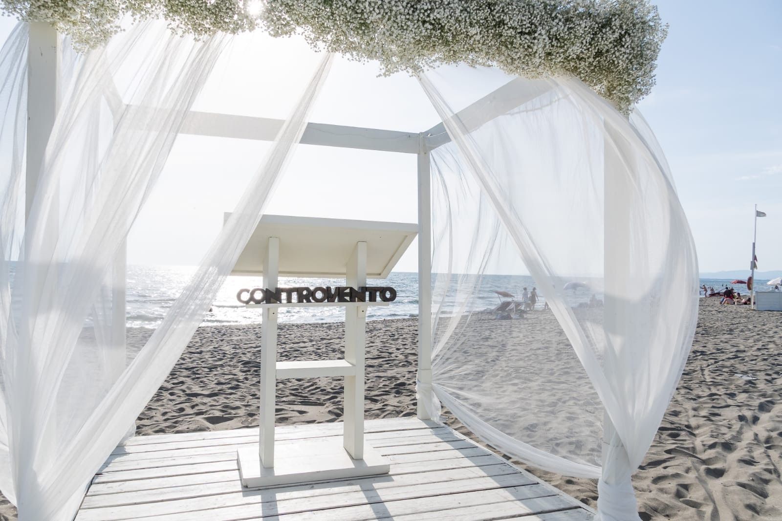 Gazebo bianco per matrimoni sulla spiaggia con tessuto bianco trasparente e dettagli floreali.