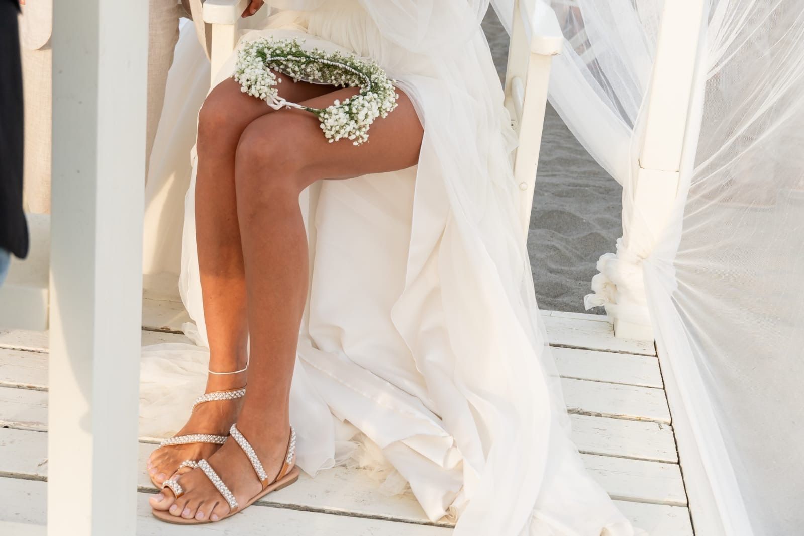Sposa in abito bianco, seduta, con fiori in mano; sandali con perline, ambiente soleggiato.