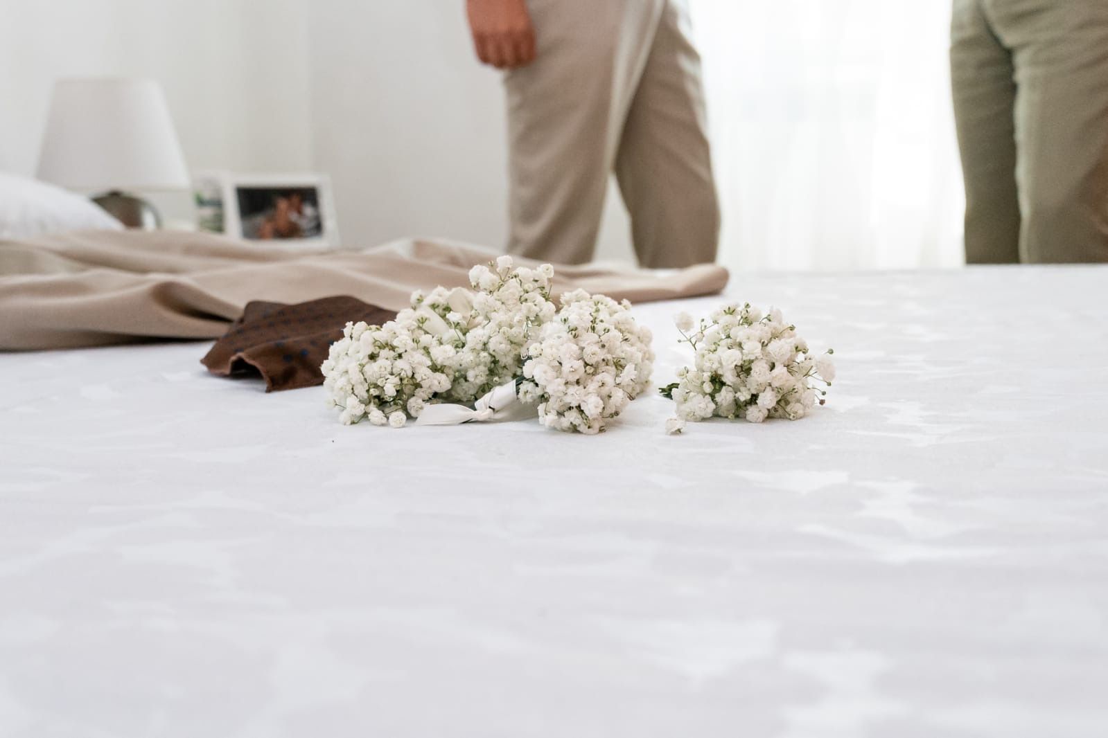 Fiori bianchi su un letto con lenzuola color cuoio. Due persone in piedi sullo sfondo.