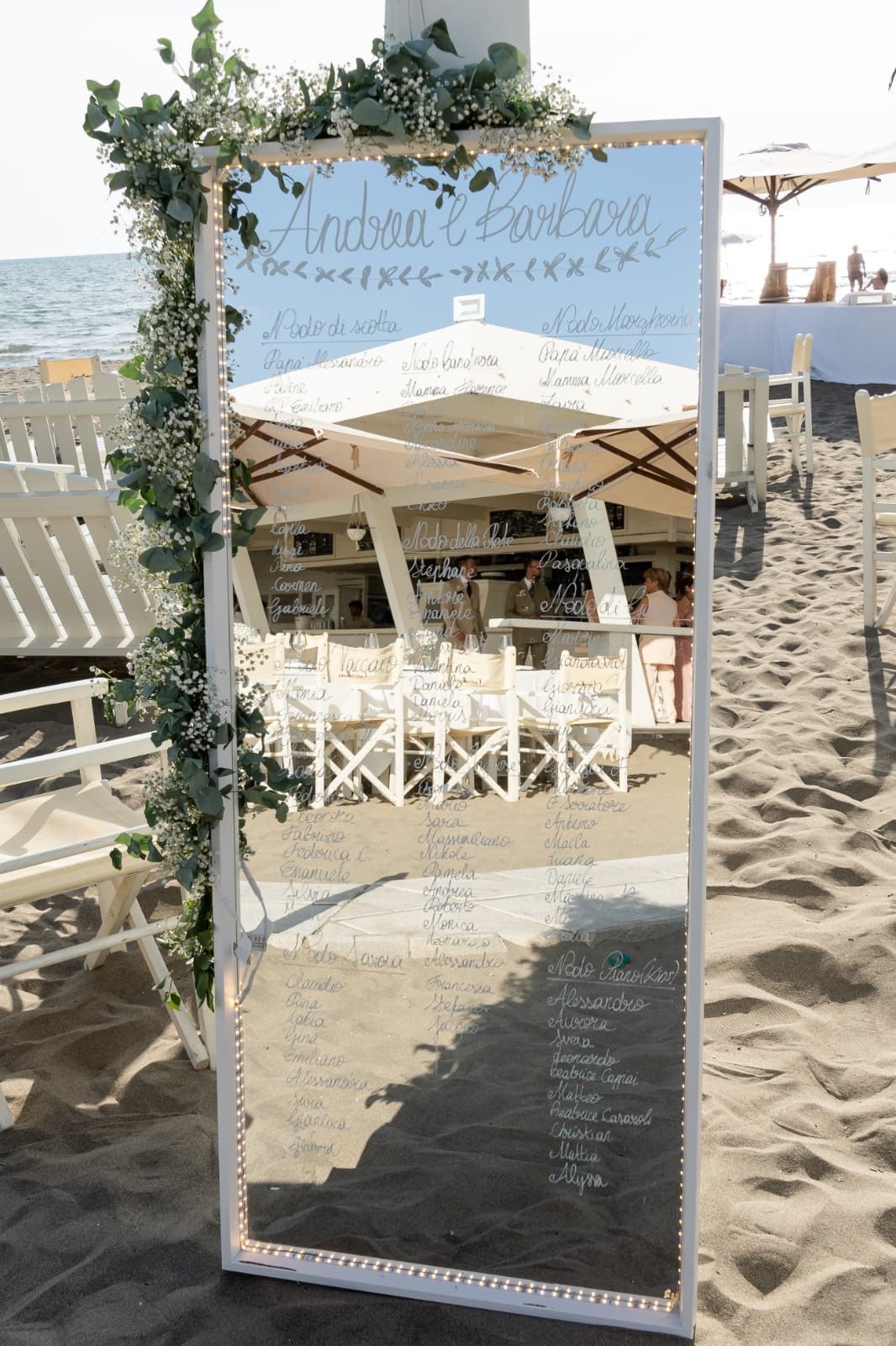 Tabella dei posti a sedere con specchio per un matrimonio in spiaggia, decorata con verde e nomi.