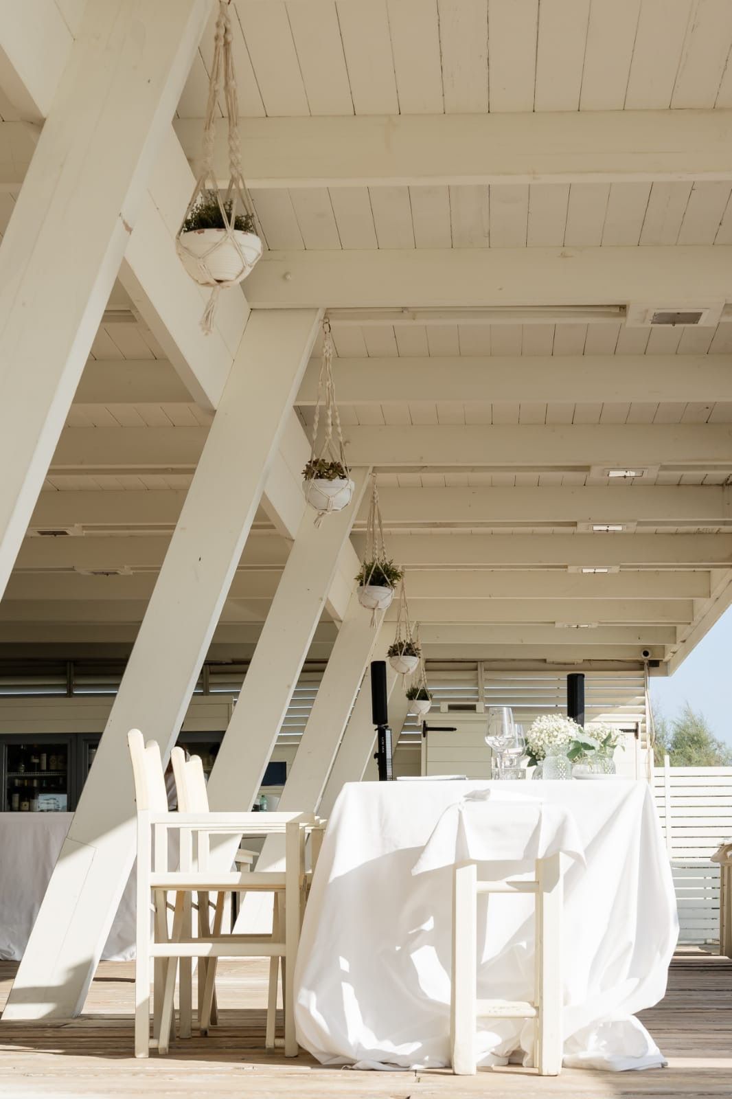 Zona pranzo all'aperto con tavoli e sedie bianchi, sotto un pergolato in legno bianco con piante pensili.