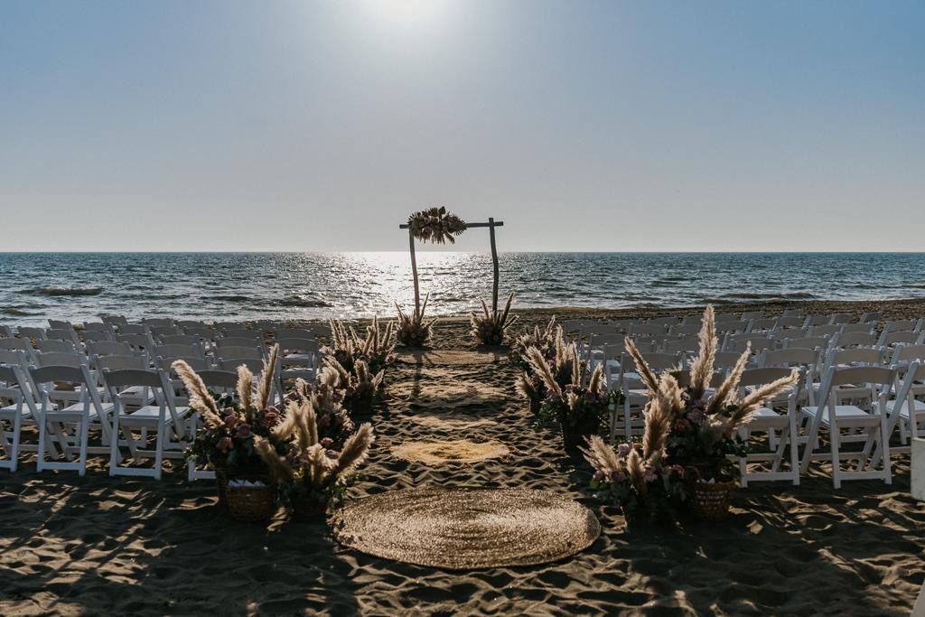 Allestimento per matrimonio in spiaggia, navata con erba della pampa che conduce a un arco di legno, sedie bianche
