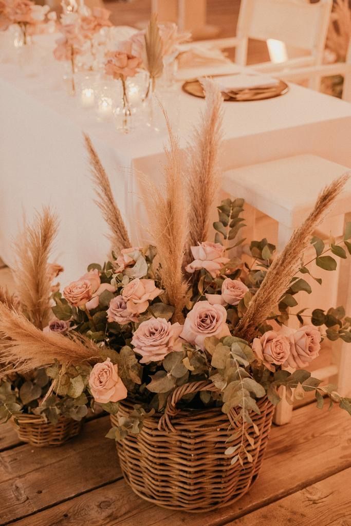 Composizione floreale in un cesto intrecciato con rose, eucalipto ed erba della pampa su un terrazzo con un tavolo bianco.
