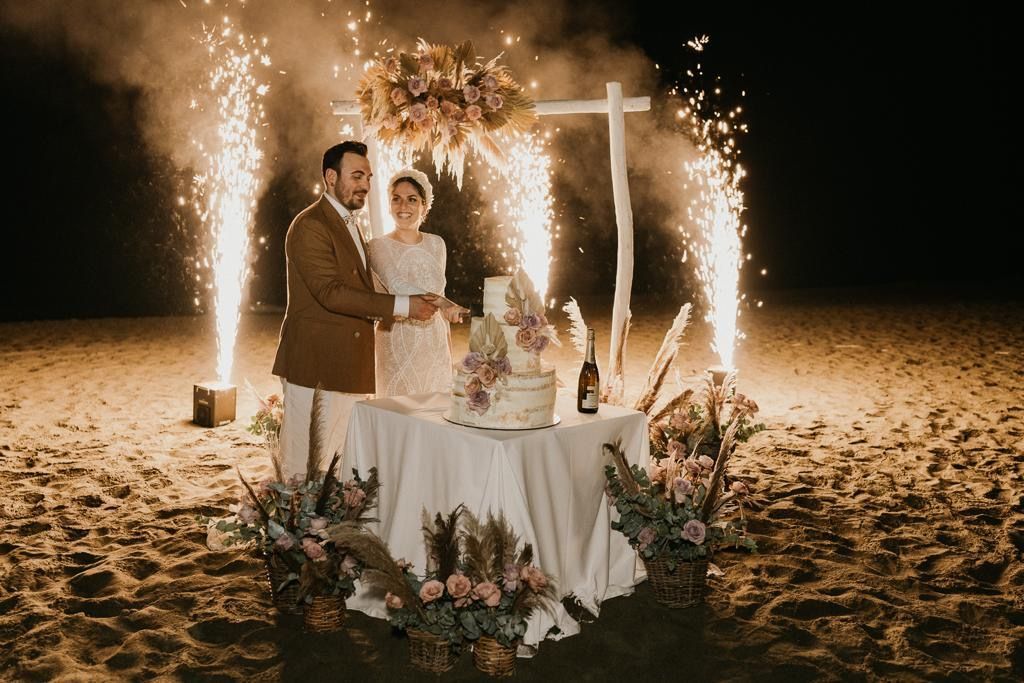 Una coppia taglia la torta a un matrimonio sulla spiaggia, con fuochi d'artificio sullo sfondo e un tavolo decorato con fiori.