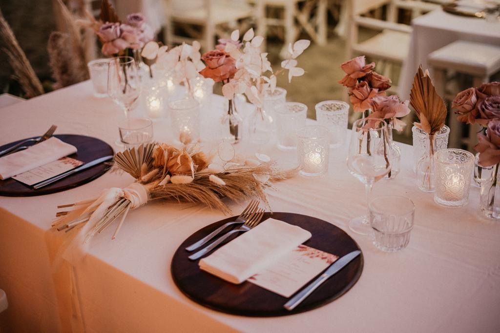 Apparecchiatura della tavola: piatti marroni, tovaglioli bianchi, fiori in vasi trasparenti, luci soffuse.