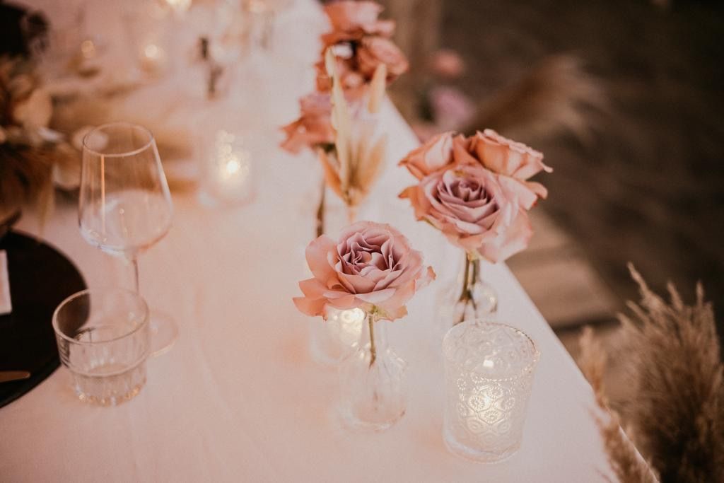 Primo piano di una tavola apparecchiata per un matrimonio con rose rosa in vasi, candele e bicchieri.