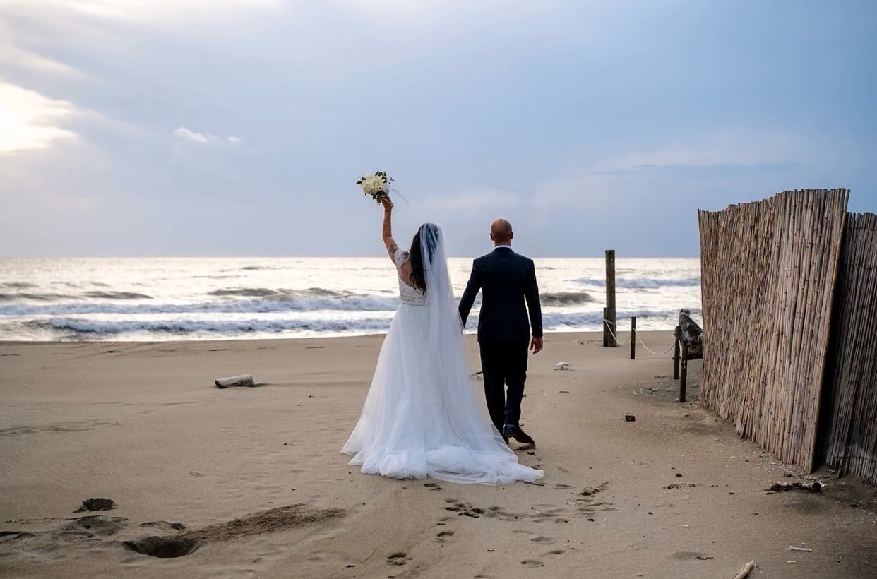 Sposi che camminano sulla spiaggia; la sposa solleva il bouquet, sullo sfondo l'oceano.