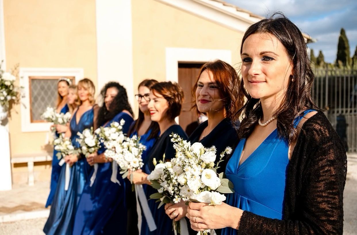 Damigelle in abiti blu con bouquet in mano, in fila fuori da un edificio.