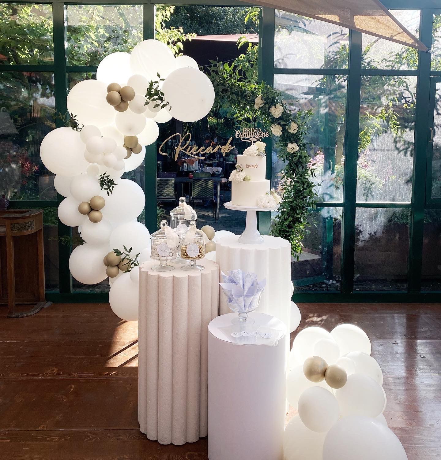 Allestimento della festa: palloncini bianchi, torta e sfondo floreale su piedistalli.