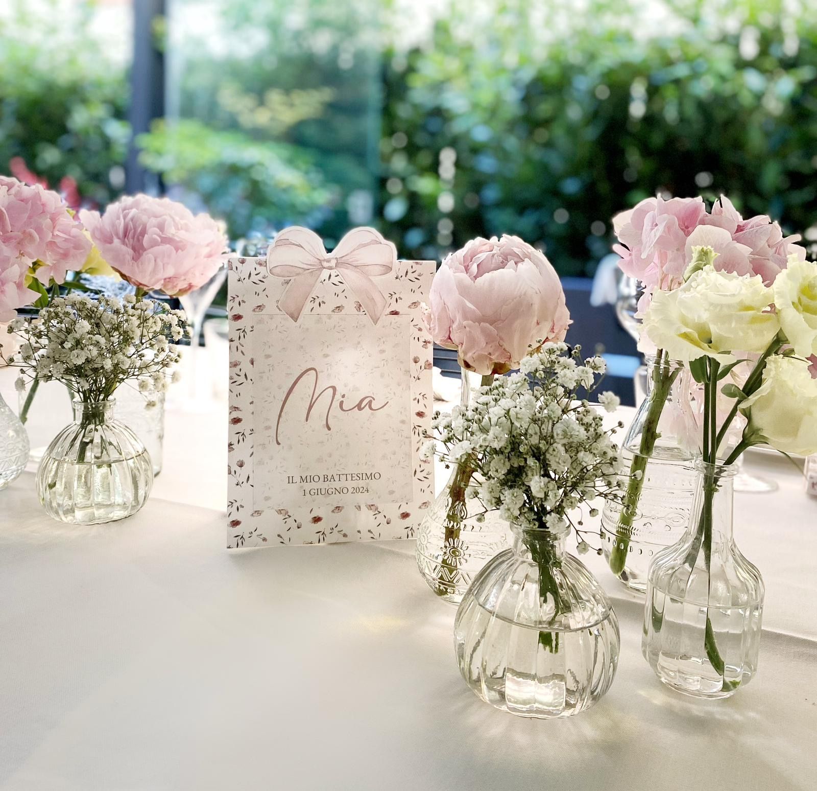 Apparecchiatura della tavola con fiori rosa, un biglietto da visita e vasi trasparenti su uno sfondo esterno sfocato.