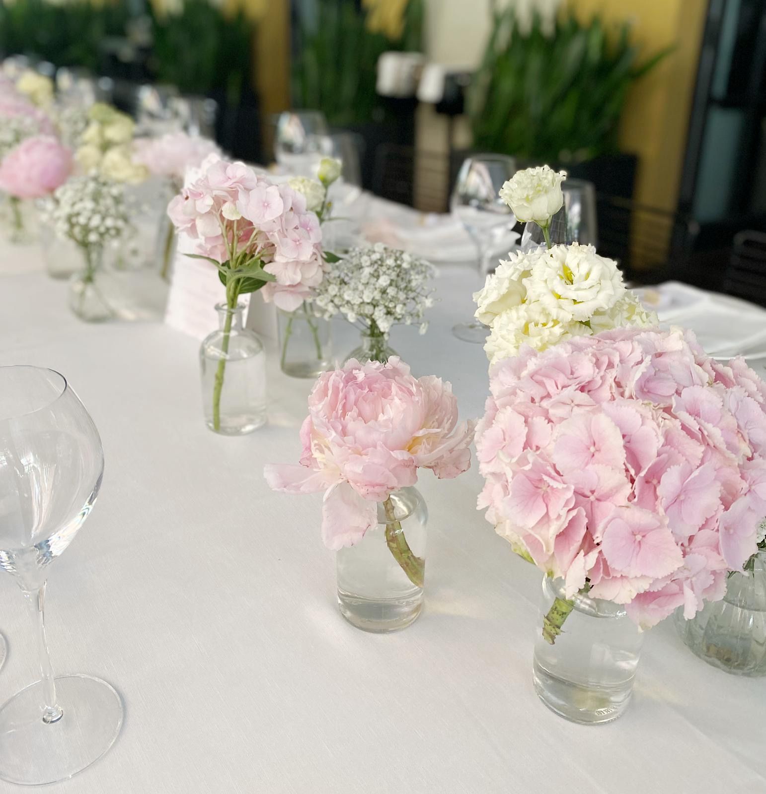 Tavolo apparecchiato con fiori rosa e bianchi in piccoli vasi di vetro, luci soffuse.