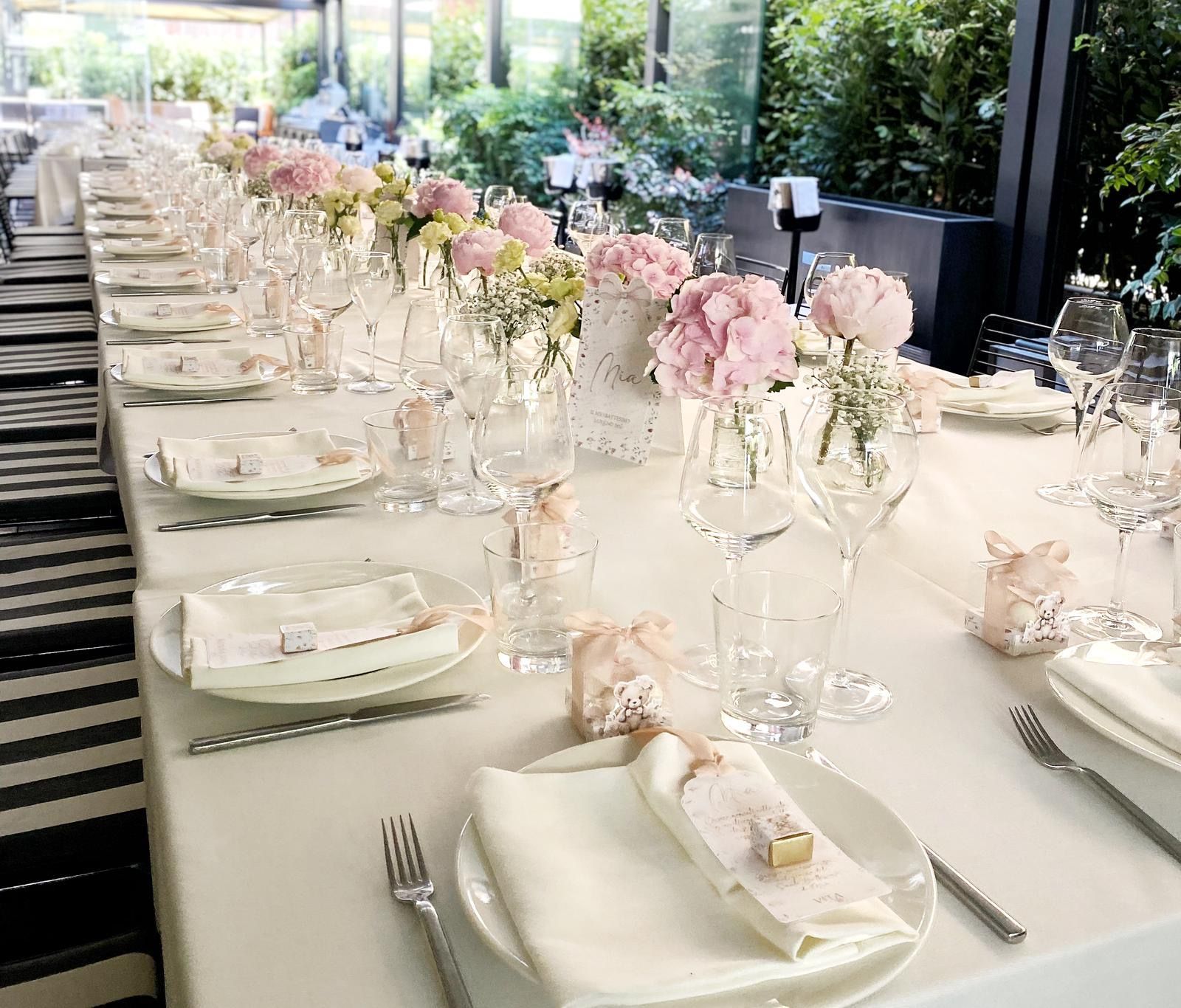 Lungo tavolo apparecchiato per un evento formale con fiori rosa, tovaglie bianche e bicchieri.