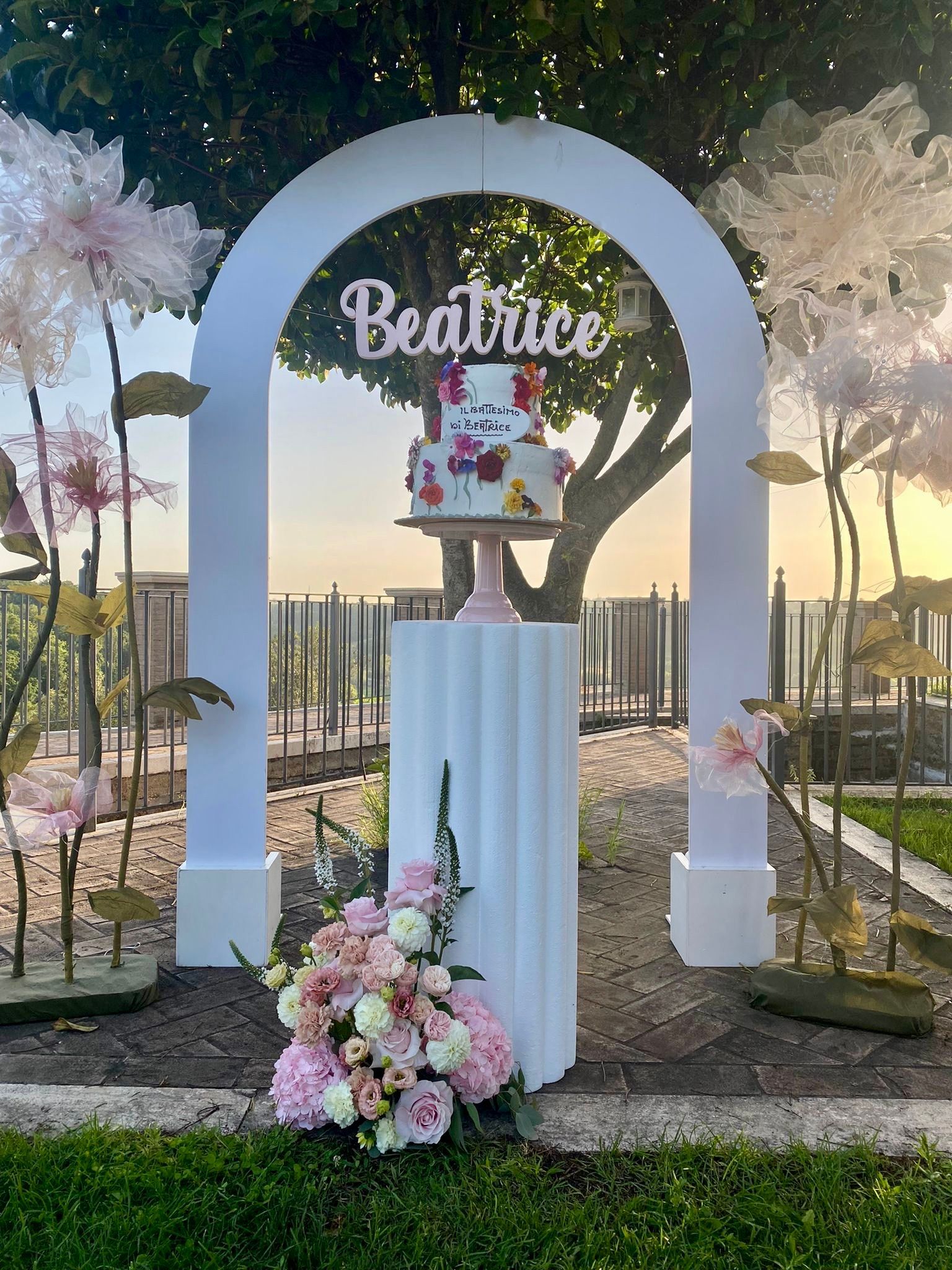La torta floreale di Beatrice è posta su un piedistallo sotto un arco bianco, con decorazioni floreali e un'ambientazione da giardino.