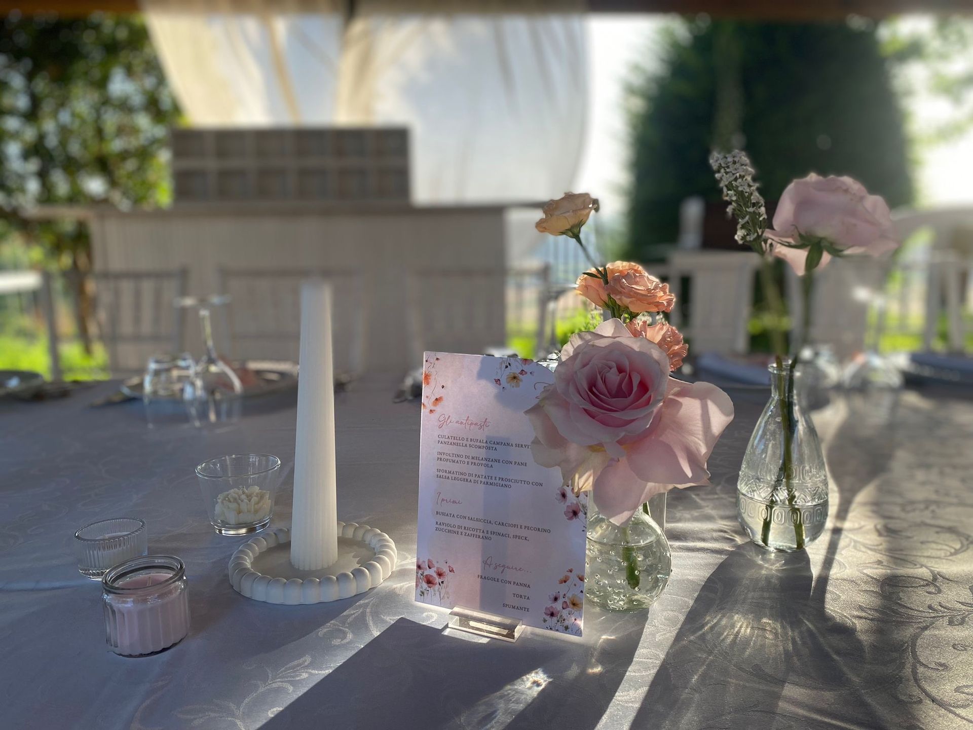 Apparecchiatura della tavola con fiori, candele e menù. Rose rosa tenui, tovaglia bianca, allestimento esterno.