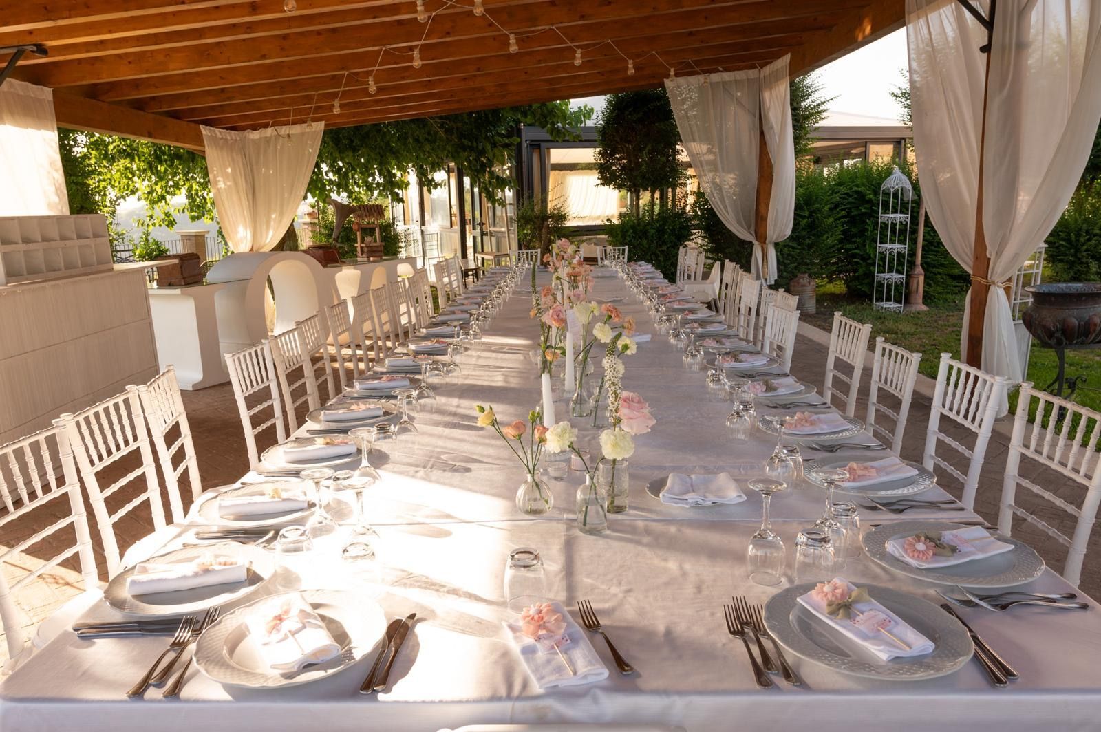 Lungo tavolo apparecchiato per un ricevimento di nozze all'aperto, sotto un pergolato in legno.