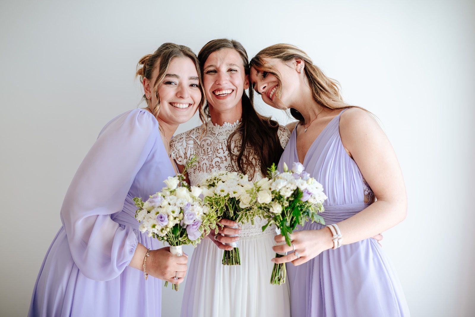 La sposa e le damigelle sorridono, tengono in mano i bouquet e indossano abiti viola chiaro e bianchi.
