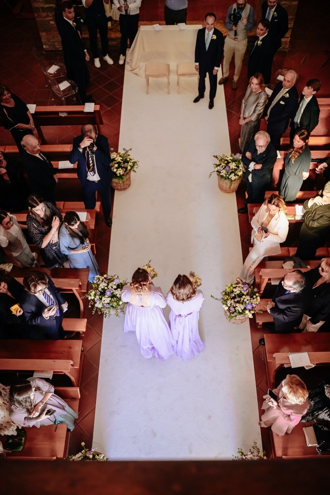 Due damigelle percorrono la navata centrale di un matrimonio verso lo sposo, con gli ospiti seduti su entrambi i lati.
