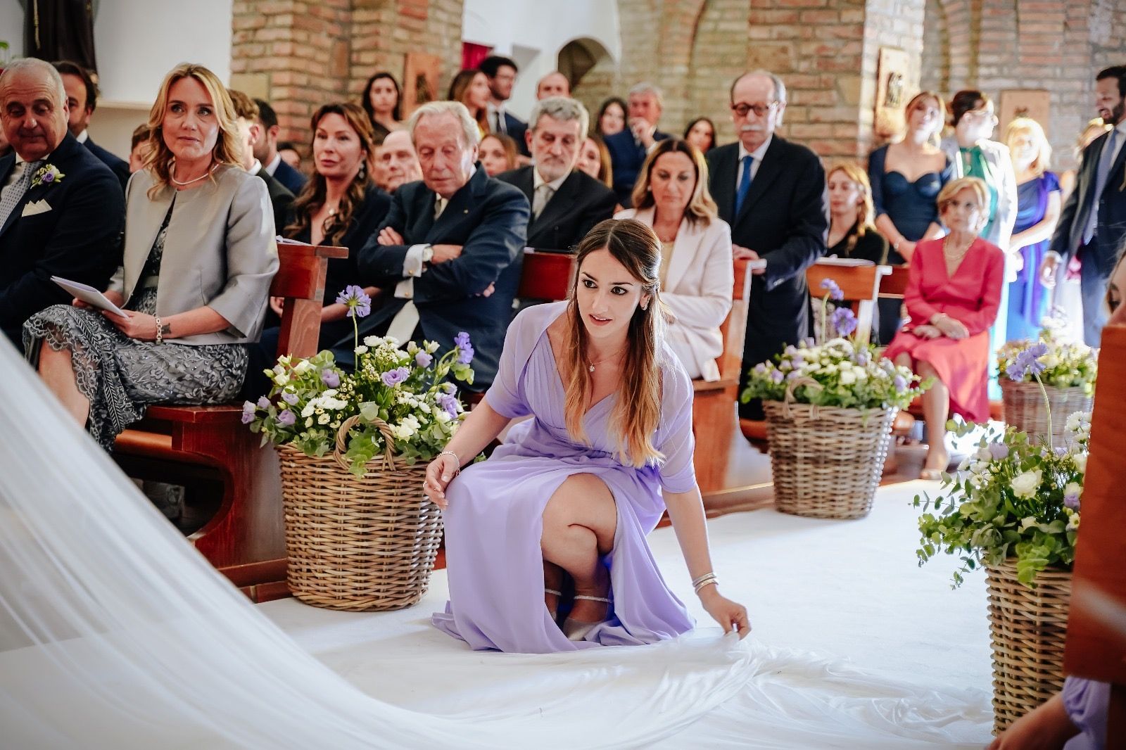 Una donna con un abito color lavanda sistema il velo da sposa nella navata, mentre gli invitati sono seduti in chiesa.