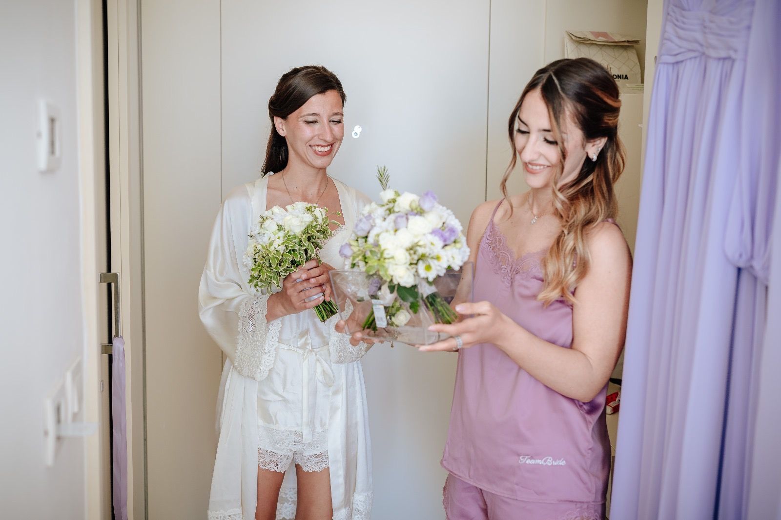 Due donne, una in vestaglia bianca e l'altra in pigiama color lavanda, sorridono in una stanza e tengono in mano dei bouquet.