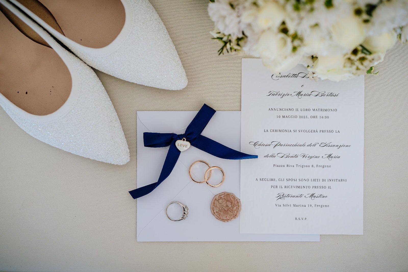 Scarpe da sposa bianche, invito con nastro blu scuro, anelli e bouquet su una superficie strutturata.