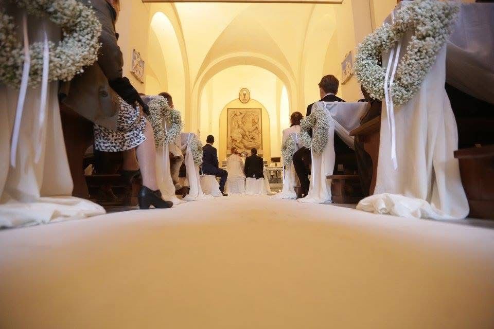 Cerimonia nuziale in chiesa; navata con tappeto bianco, decorazioni floreali e persone sedute.
