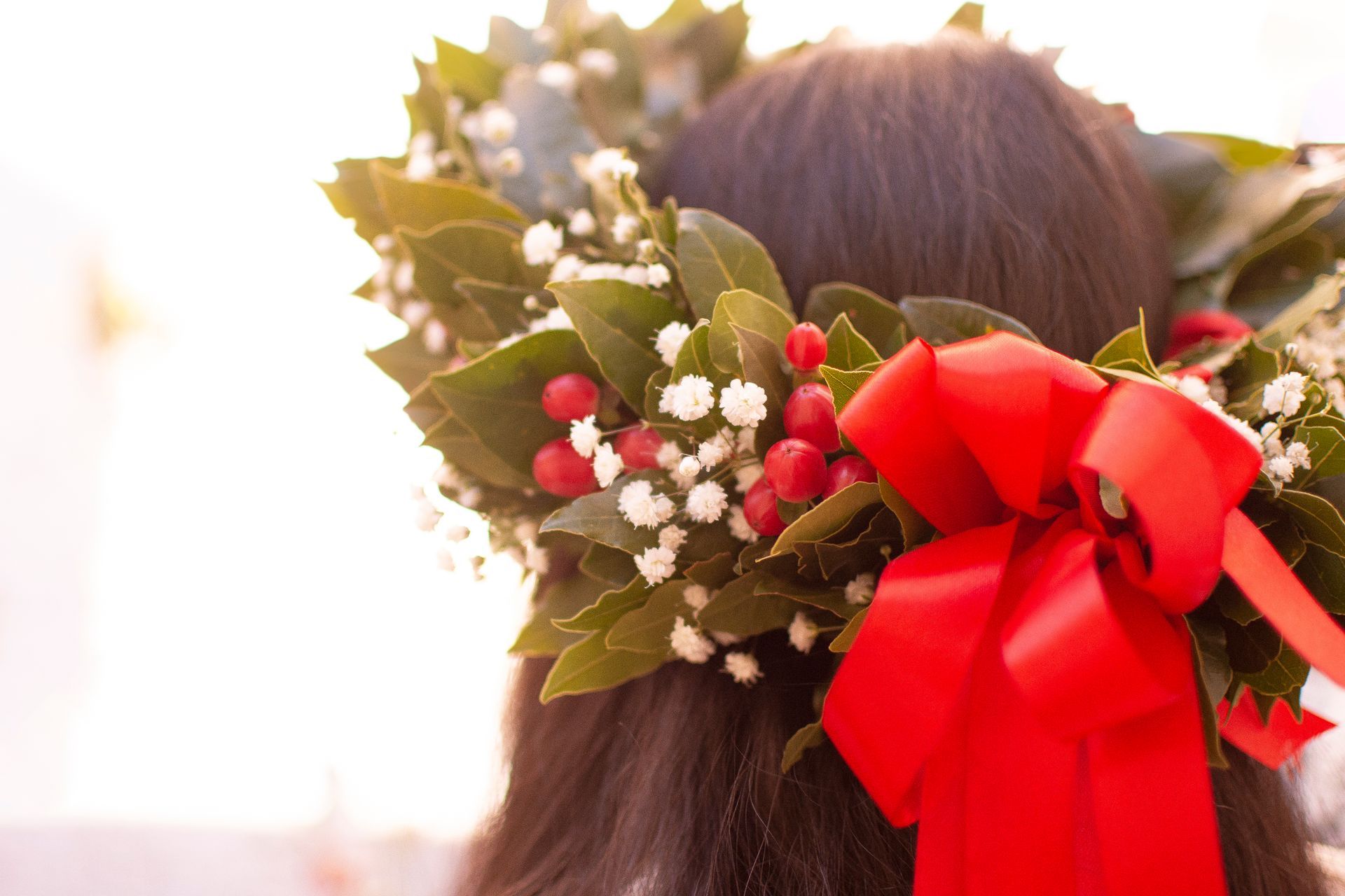 Capelli scuri con corona floreale di foglie, fiori bianchi e bacche rosse, legata con un fiocco rosso.