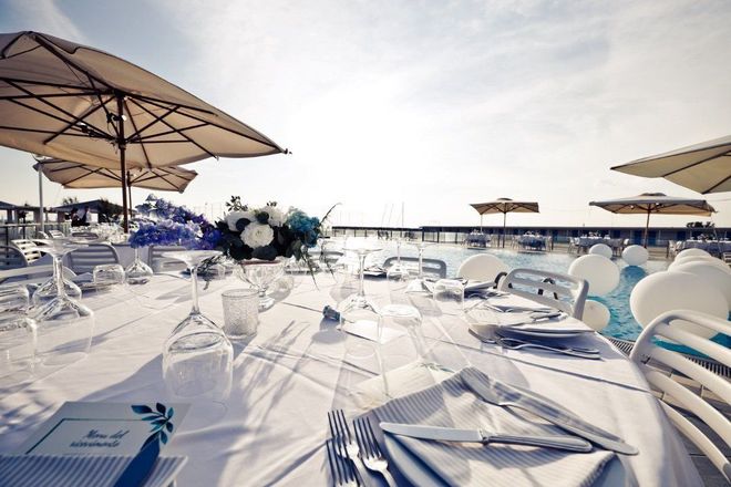 Tavolo apparecchiato per un evento all'aperto con tovaglia bianca, composizione floreale e ombrelloni sopra la testa.