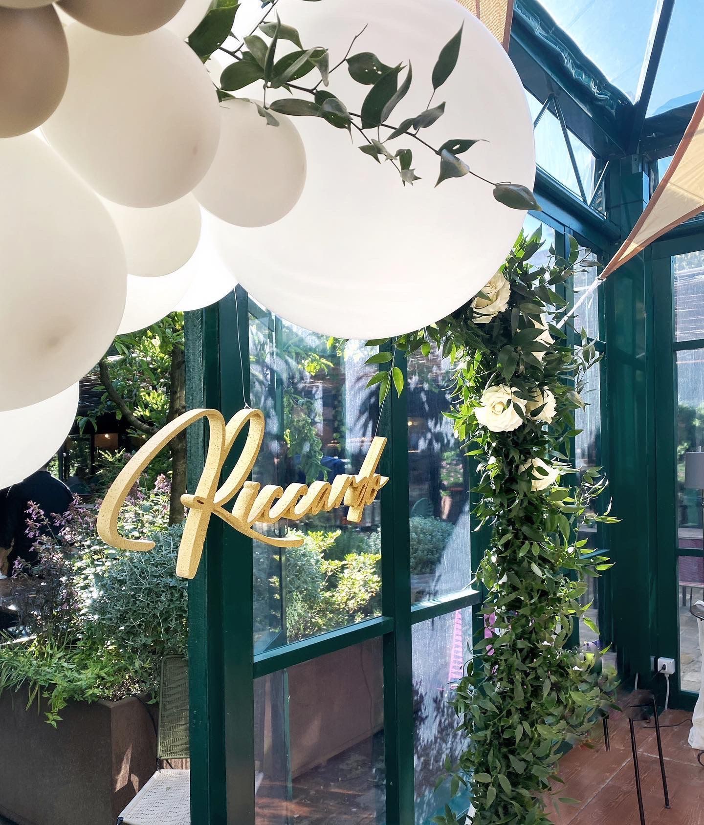 Decorazioni per feste con palloncini, verde e scritte dorate in un ambiente interno illuminato dal sole.