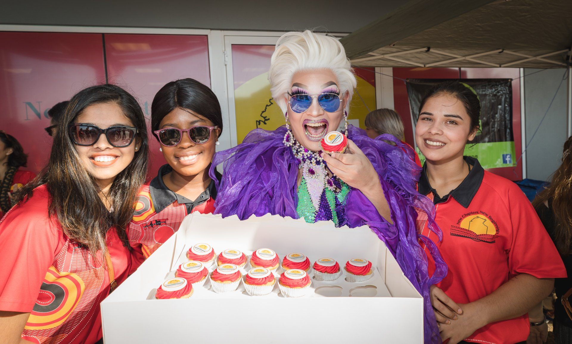 A group of people are standing around a box of cupcakes.