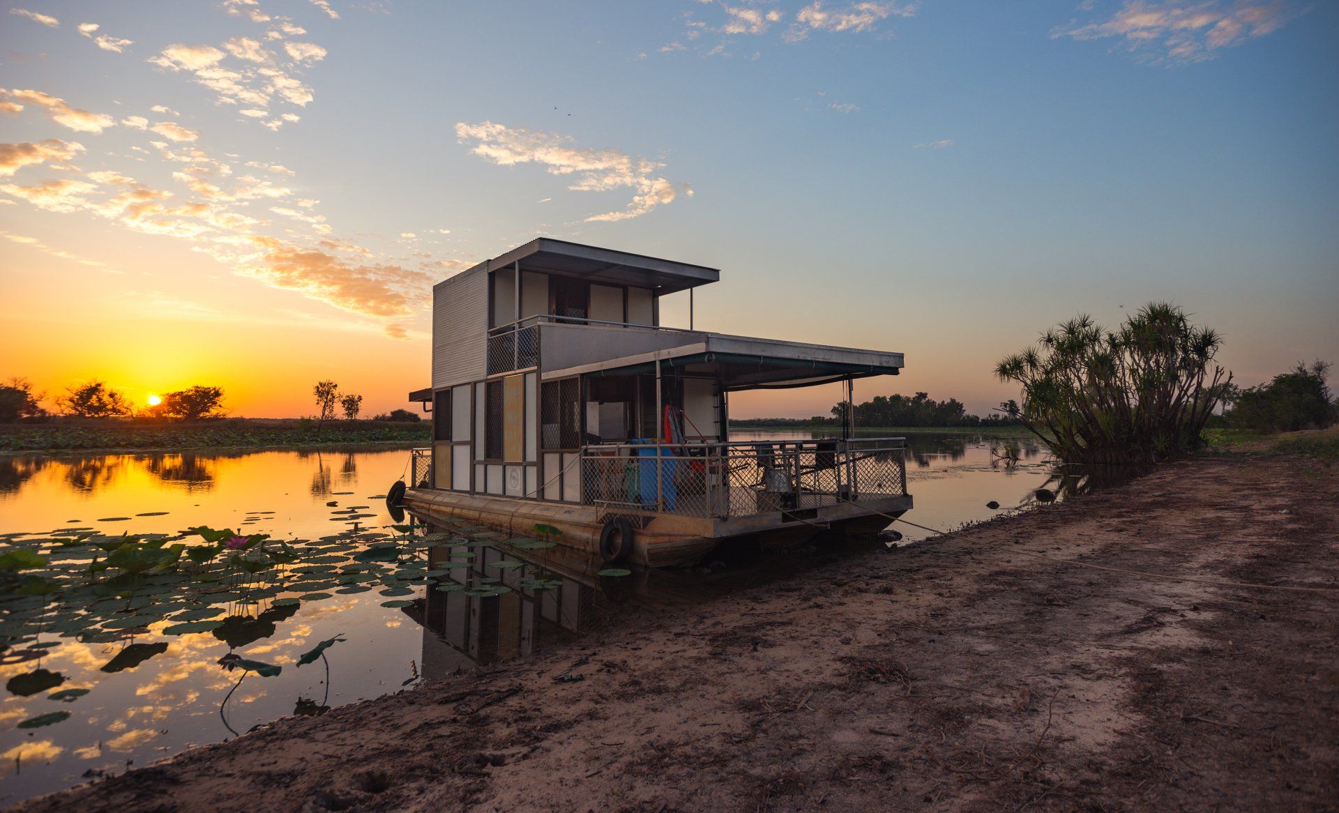 A floating house is sitting on the shore of a lake at sunset.