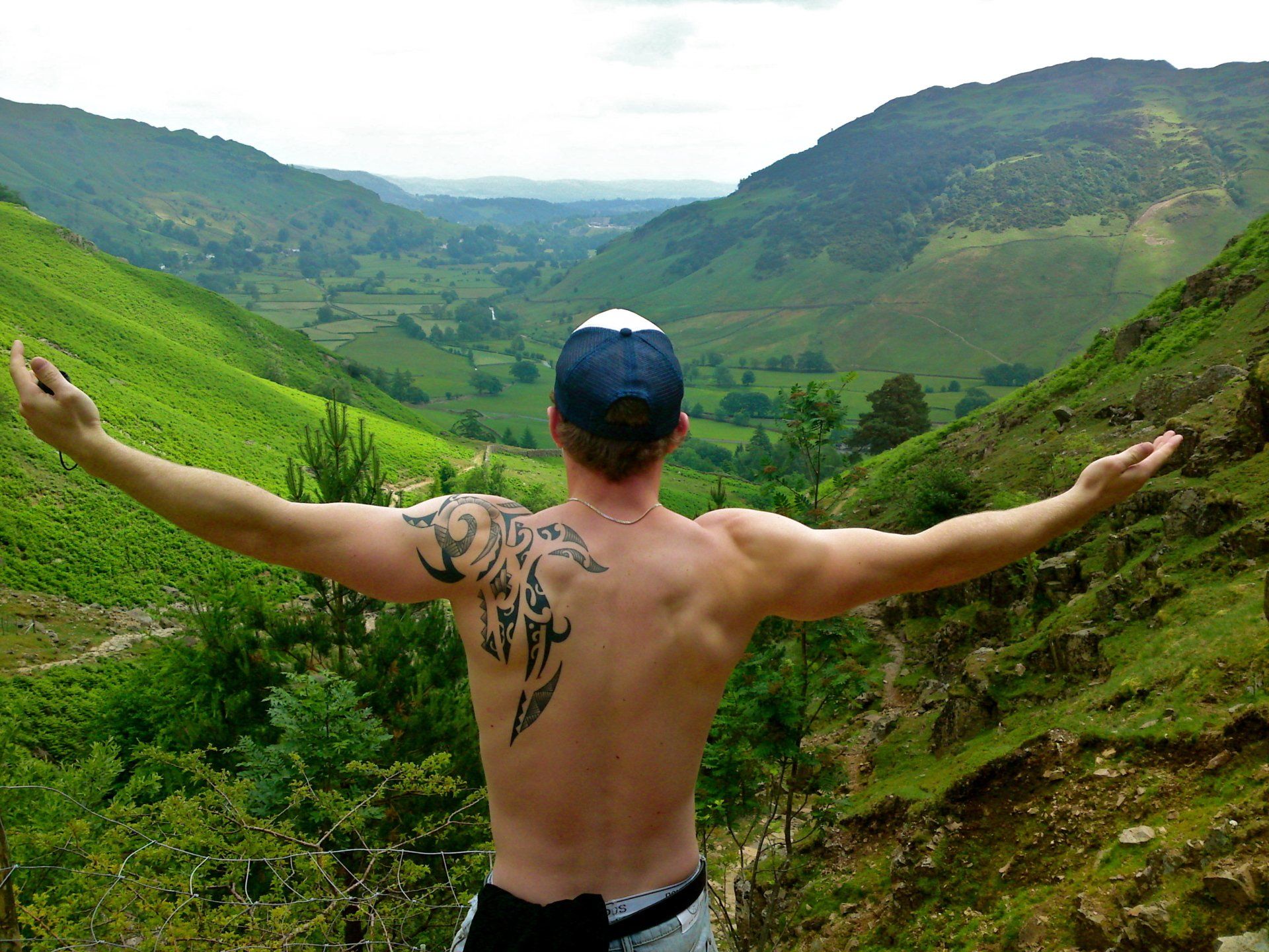 A shirtless man with a tattoo on his back is standing on top of a hill with his arms outstretched