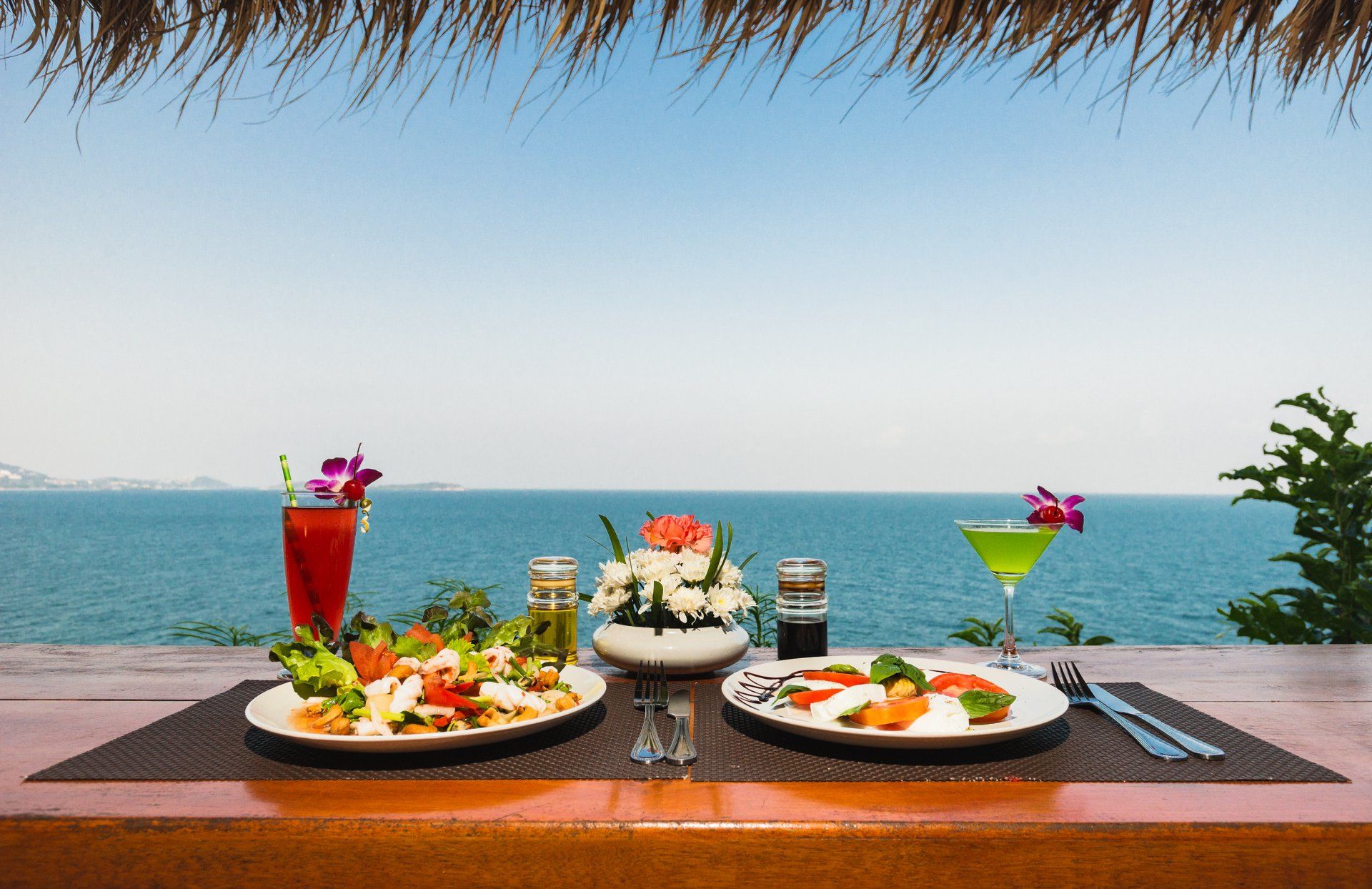 Two plates of food and two drinks on a table with a view of the ocean.