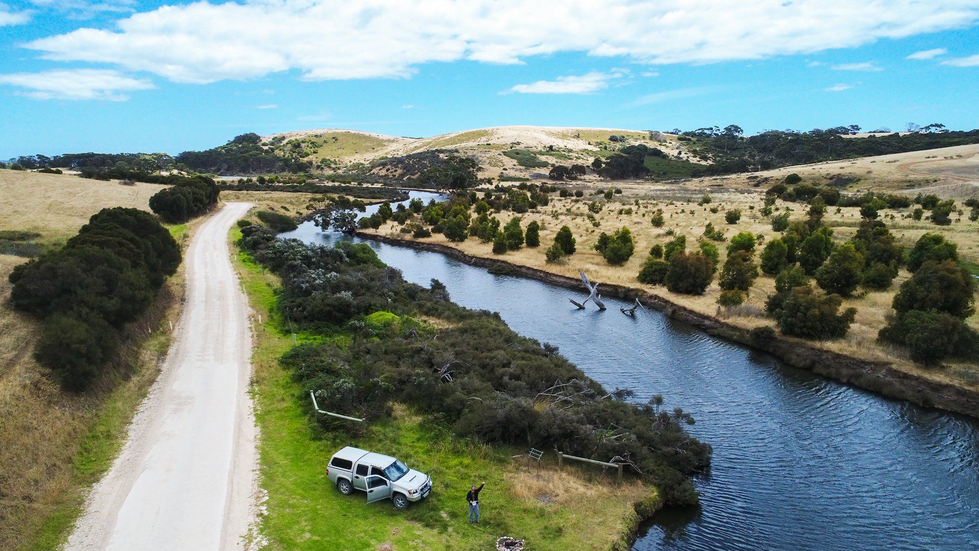 An aerial view of a car parked on the side of a dirt road next to a river.