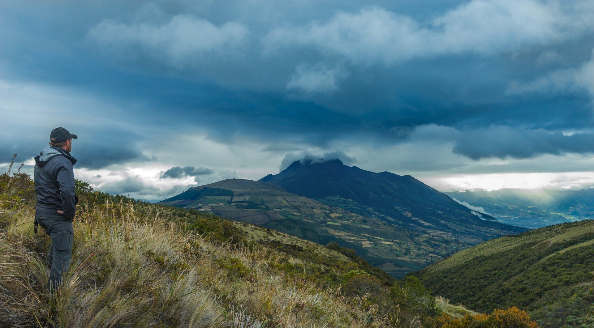 A man is standing on top of a hill looking at a mountain.