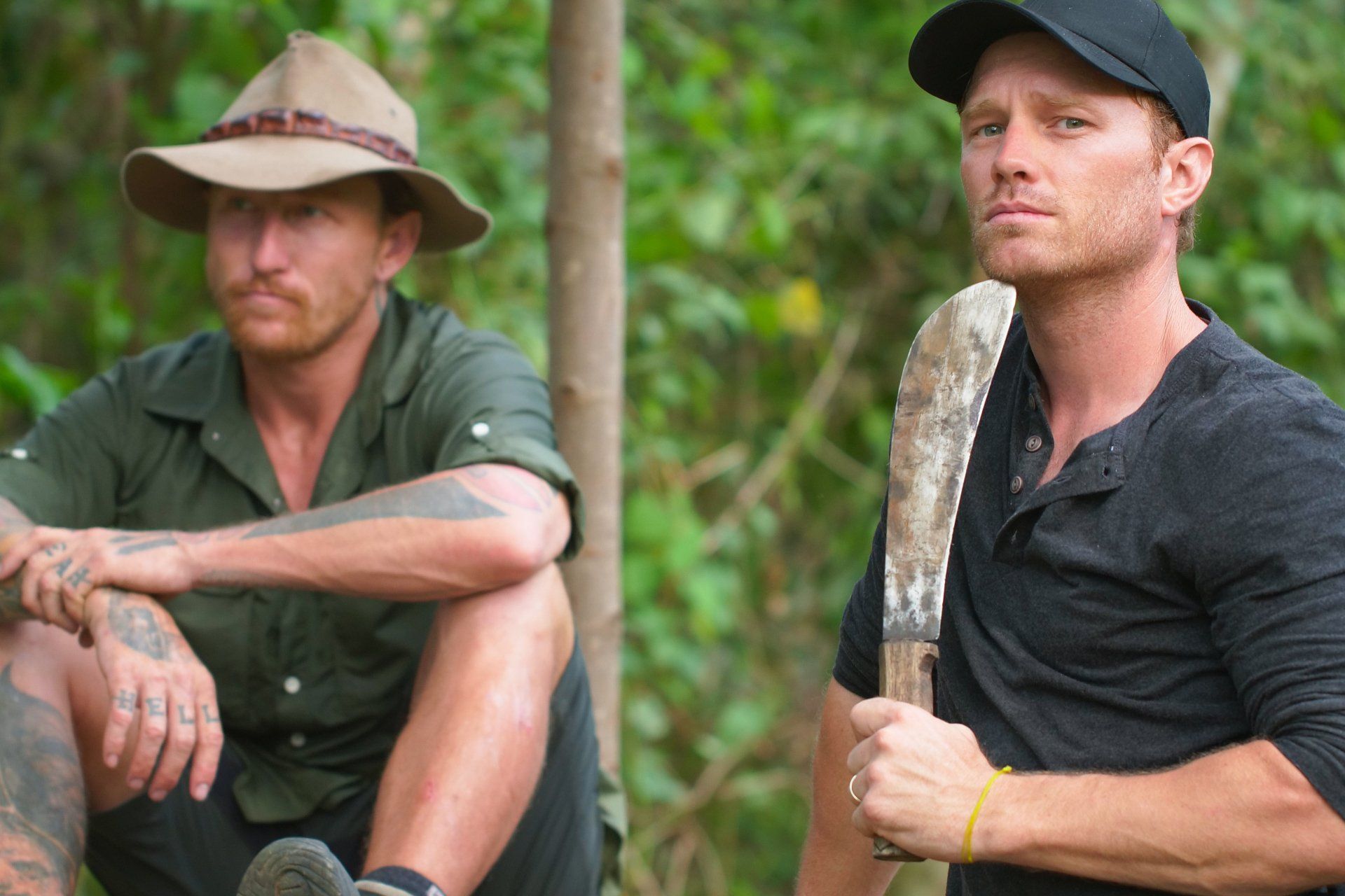 Two men are sitting next to each other in the woods . one of the men is holding a machete.
