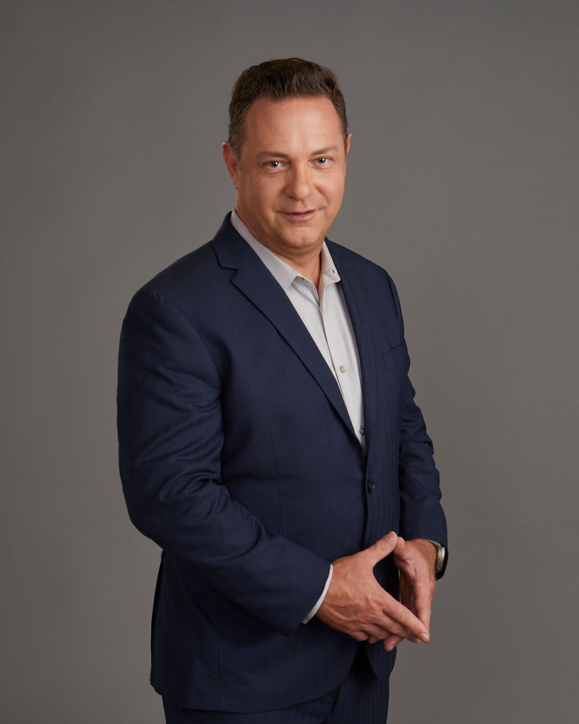 Man in navy suit, hands clasped, smiling, gray background.