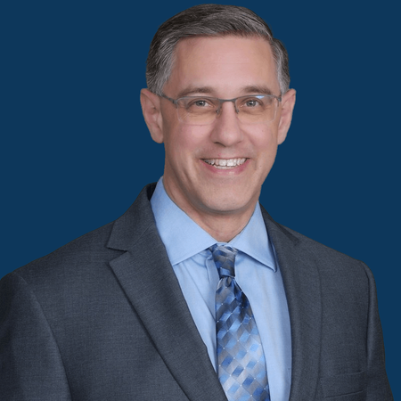 Man in suit, smiling, wearing glasses. Blue tie, light blue shirt, blue background.