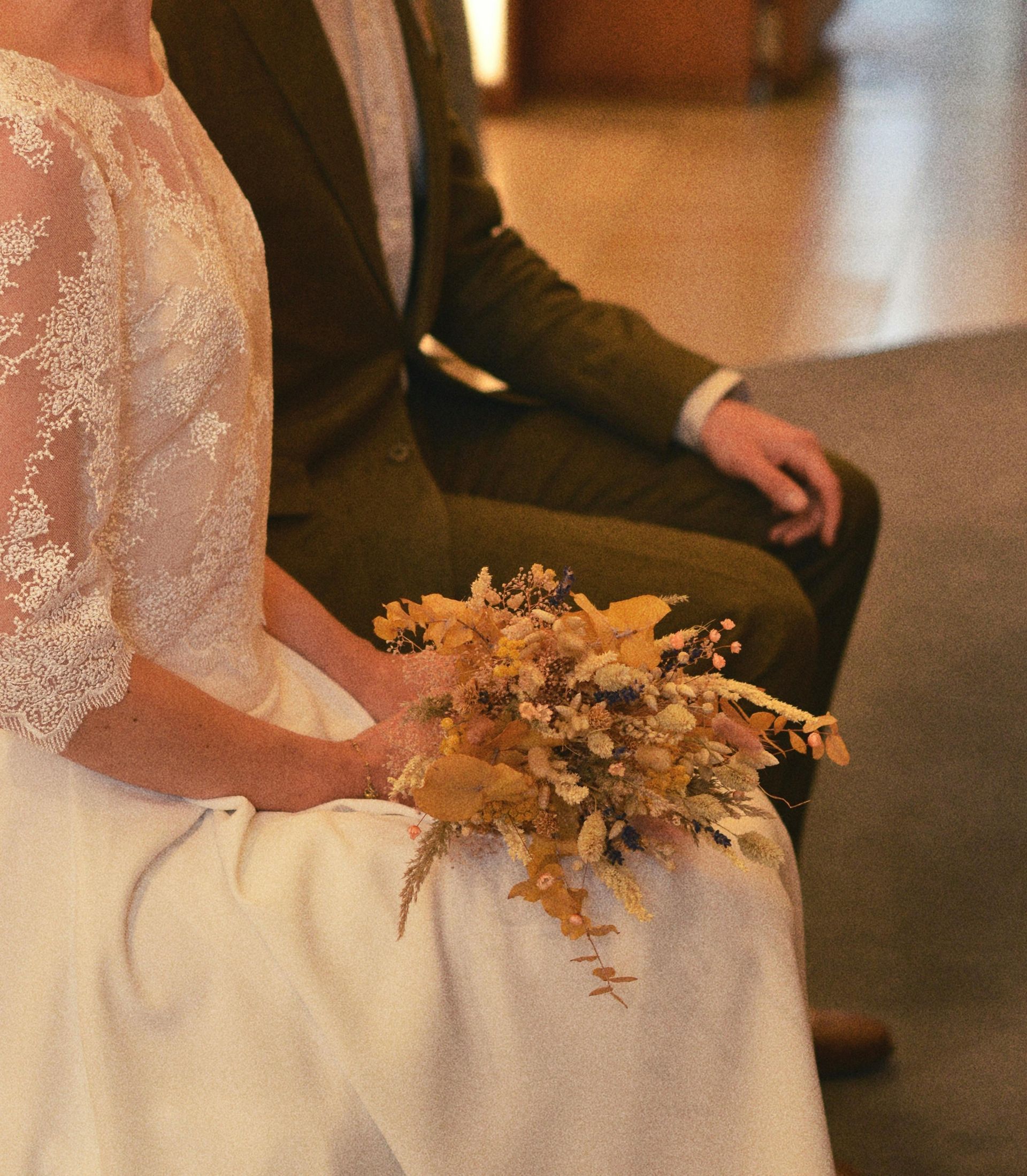 A person in a lace wedding dress holds a dried flower bouquet while sitting next to a person in an olive suit.
