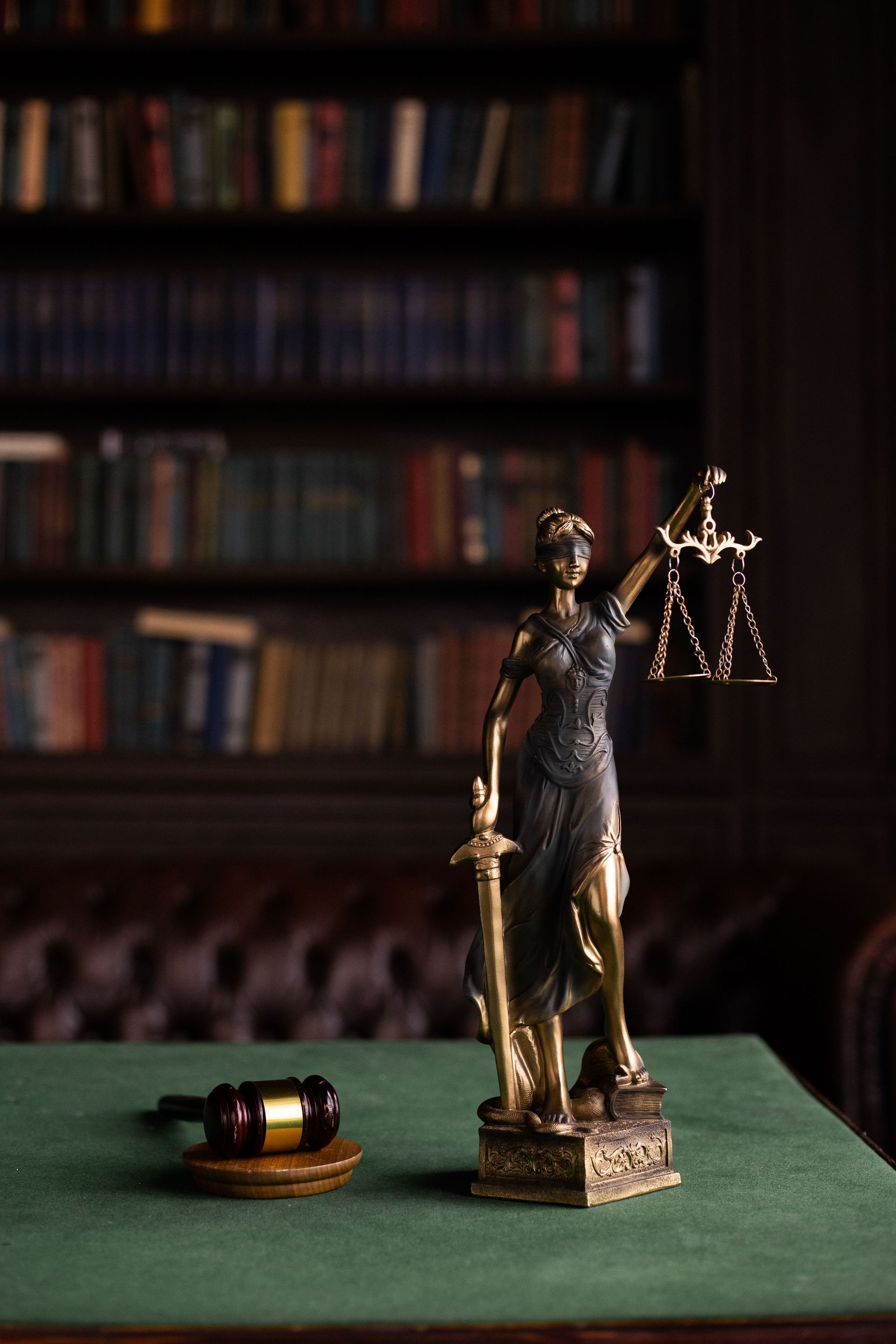 Lady Justice statue on a green desk, with scales and a gavel in front of a blurred bookshelf backdrop