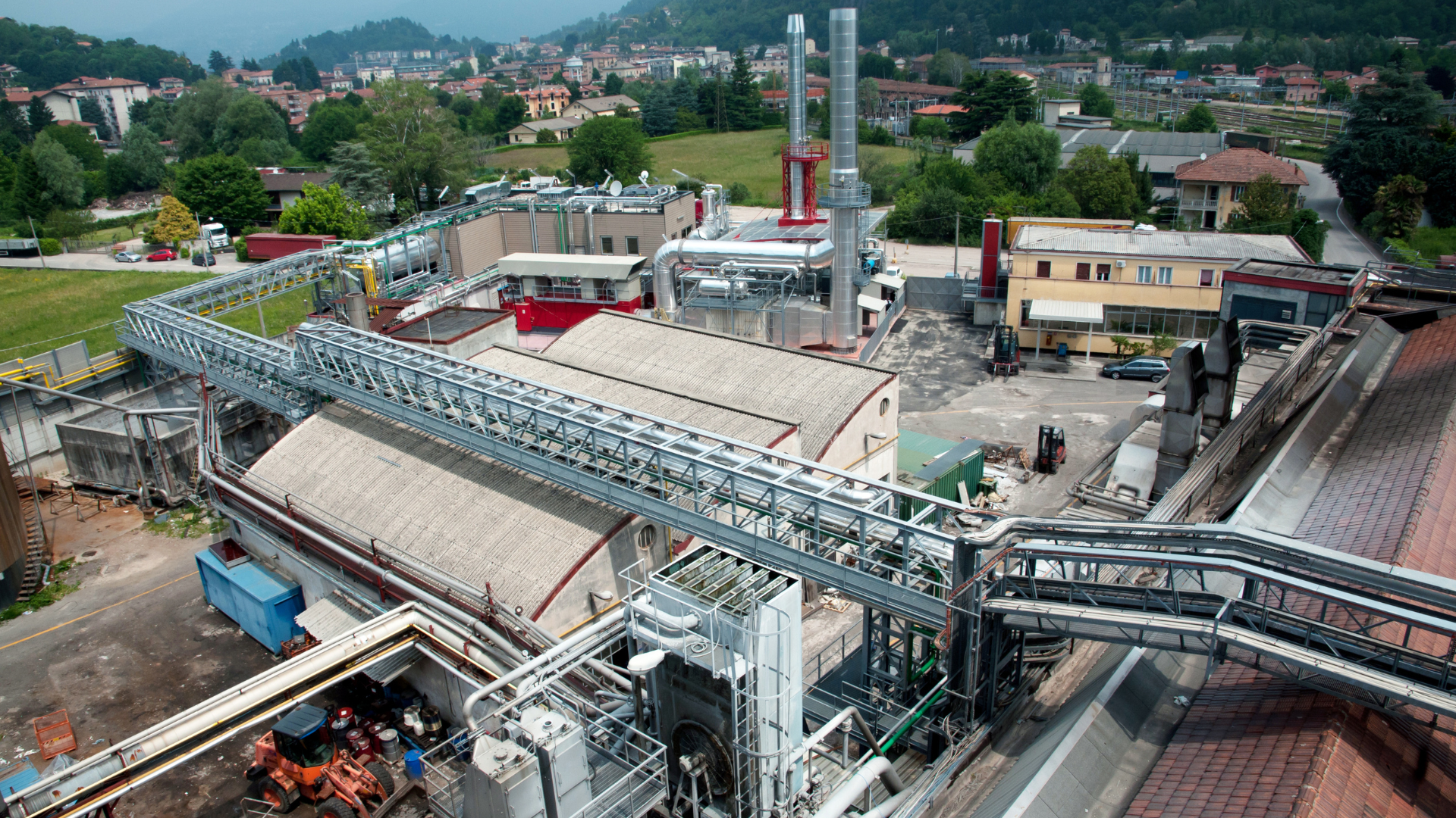 Vista aérea de una instalación industrial con edificios, tuberías, chimeneas y una cinta transportadora, ubicada en una ciudad con colinas verdes.