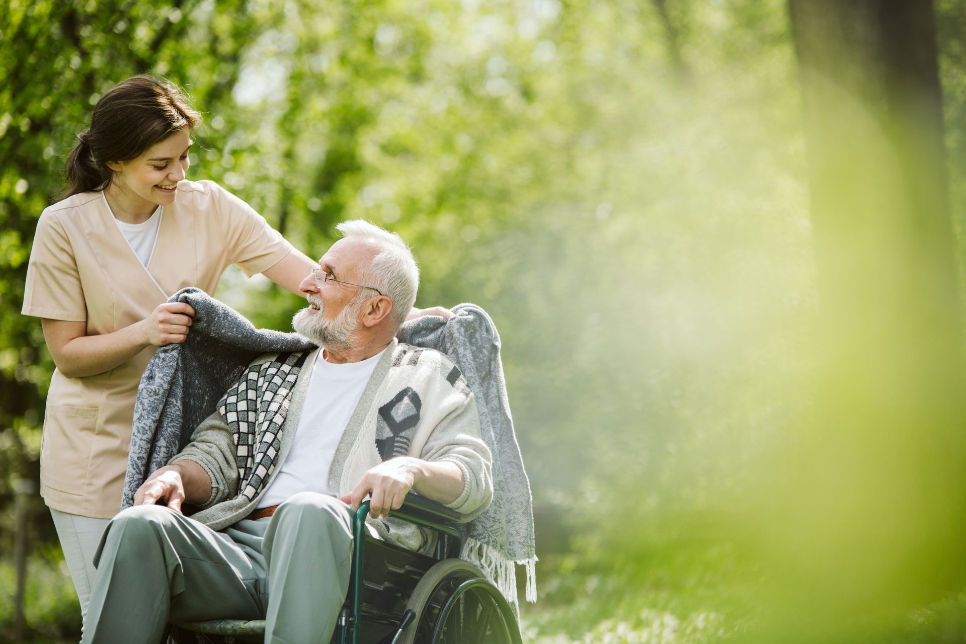 Woman helps person in wheelchair with a blanket outside; sunny day, green background.