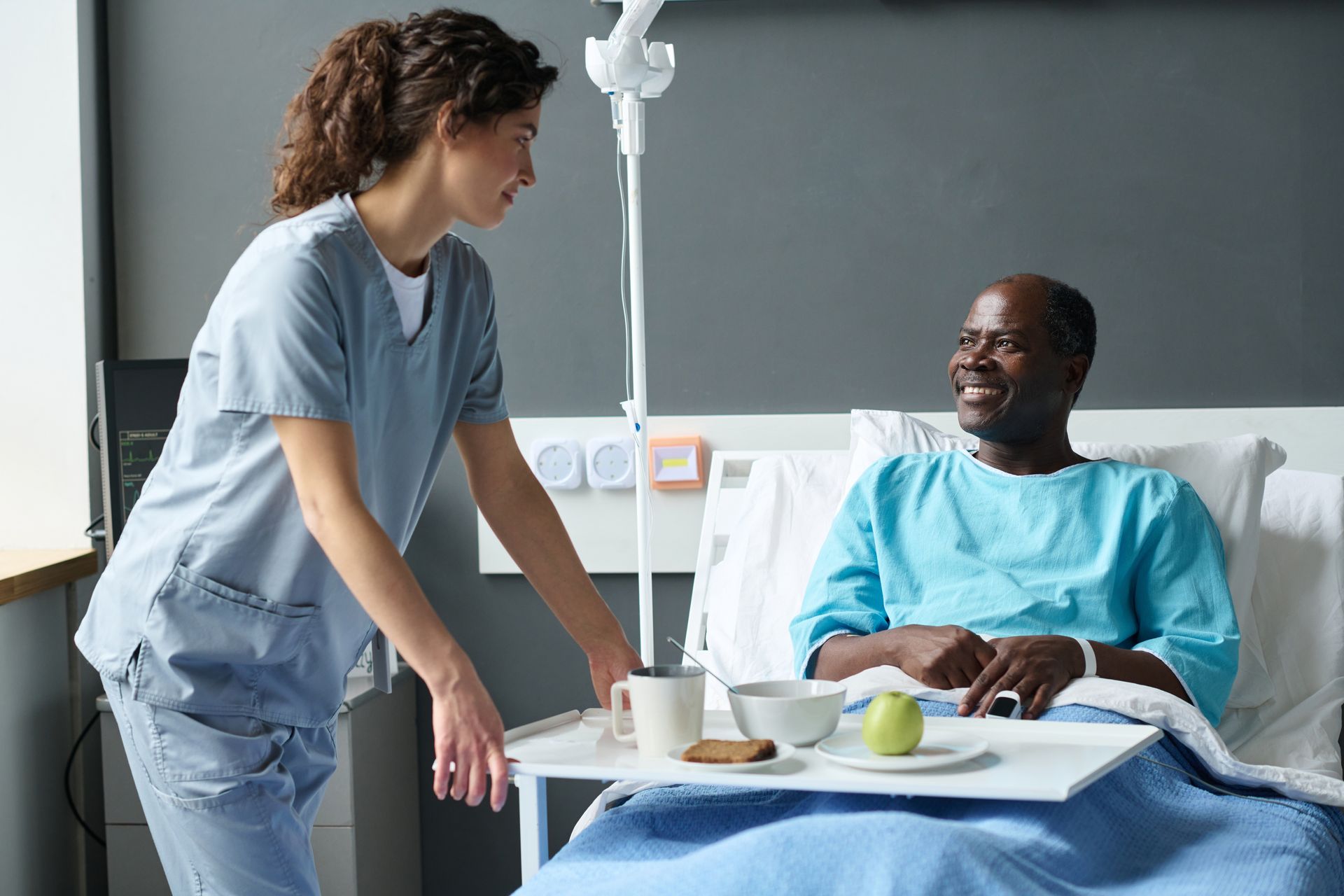 Nurse delivering food tray to smiling patient in hospital bed.