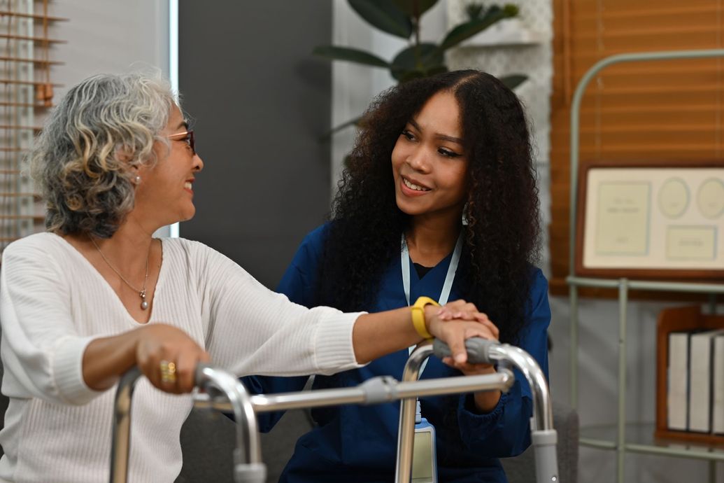 Woman helps another woman with a walker. They're in an indoor setting, smiling.
