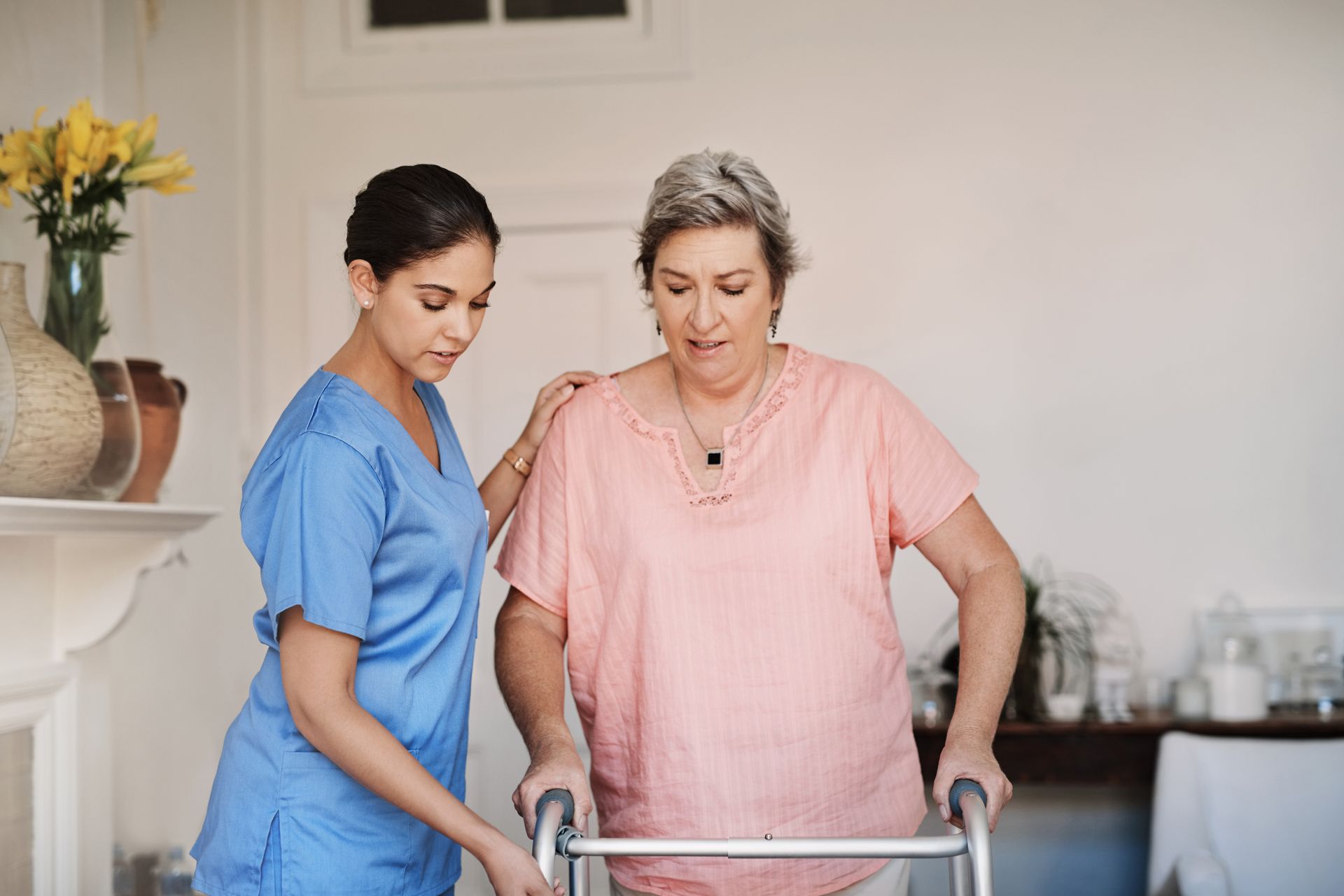 Caregiver assists a person using a walker, indoors. Light-skinned person with short gray hair. Caregiver in blue scrubs.