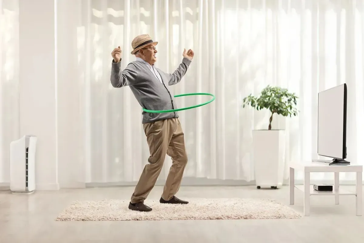 Senior man hula hooping on top of a carpet flooring in his living room.