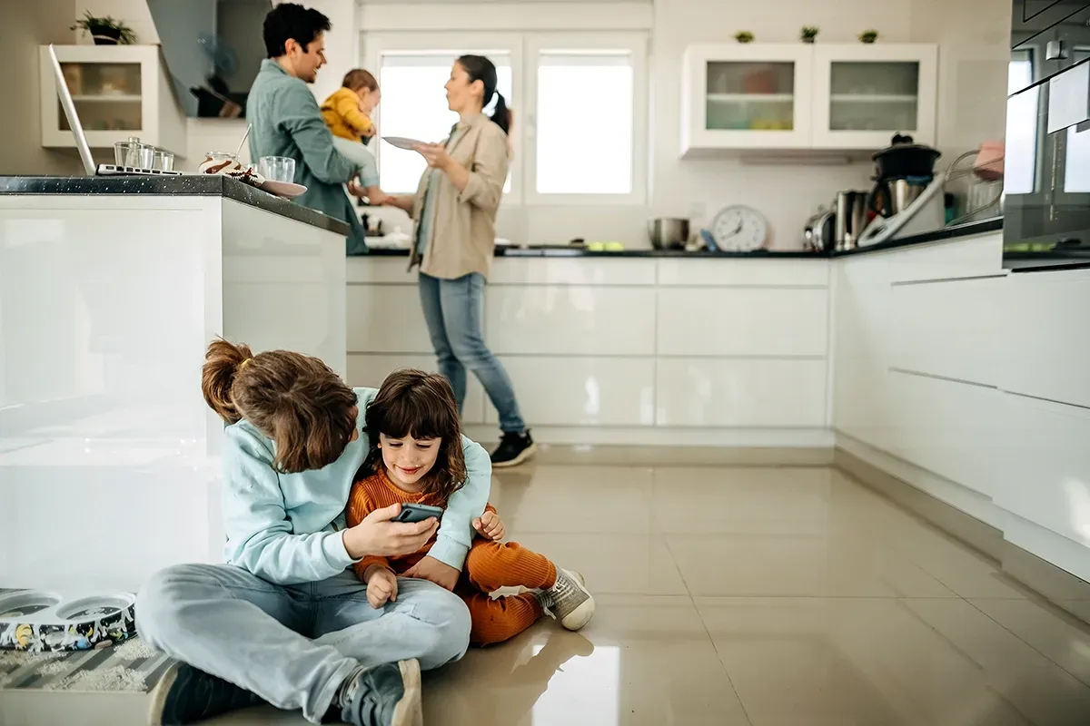 Kids sitting on the kitchen floor with parents holding a baby in the background.
