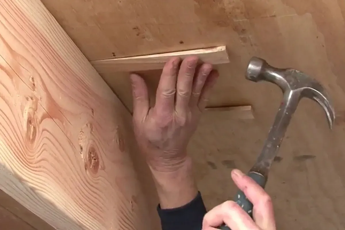 Close up of a hammer tapping in a wood shim between the subfloor and joint to stabilize a squeaky floor.