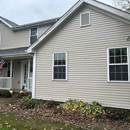 Two-story beige house with white trim and windows. Porch with American flag. Overcast day with fall foliage.