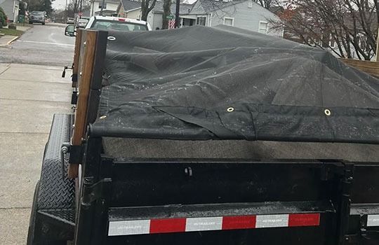 Black tarp covers a trailer bed, secured by wooden posts on the side. Reflective strip at the back.