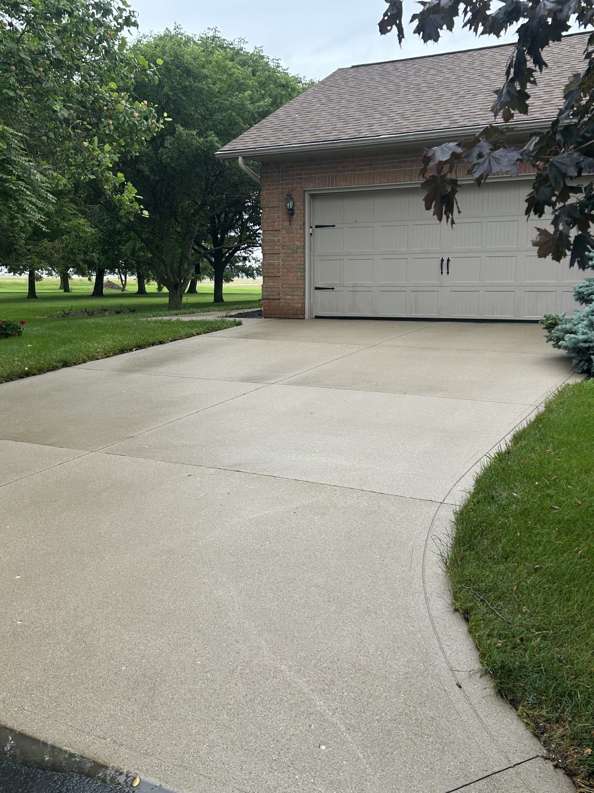 Concrete driveway leads to a beige garage with a brown tiled roof, surrounded by green grass and trees.